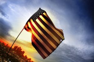 Wide angle photo of a tattered American flag blowing in the wind over a distant city lit by golden sunset light